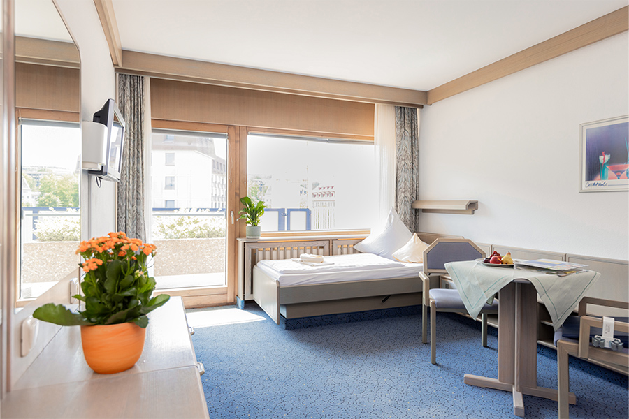 Ein beispielhaftes Patientenzimmer mit großen Fenstern, Balkon, Fernseher, Bett und Sitzecke.