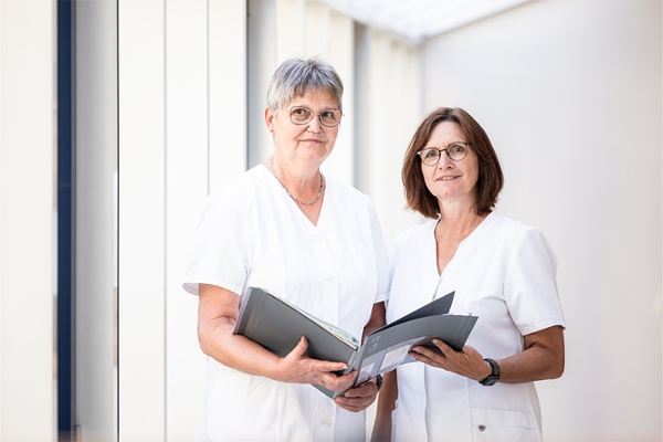 Zwei Frauen vom Klinikpersonal halten eine Patientenakte und lächeln in die Kamera.