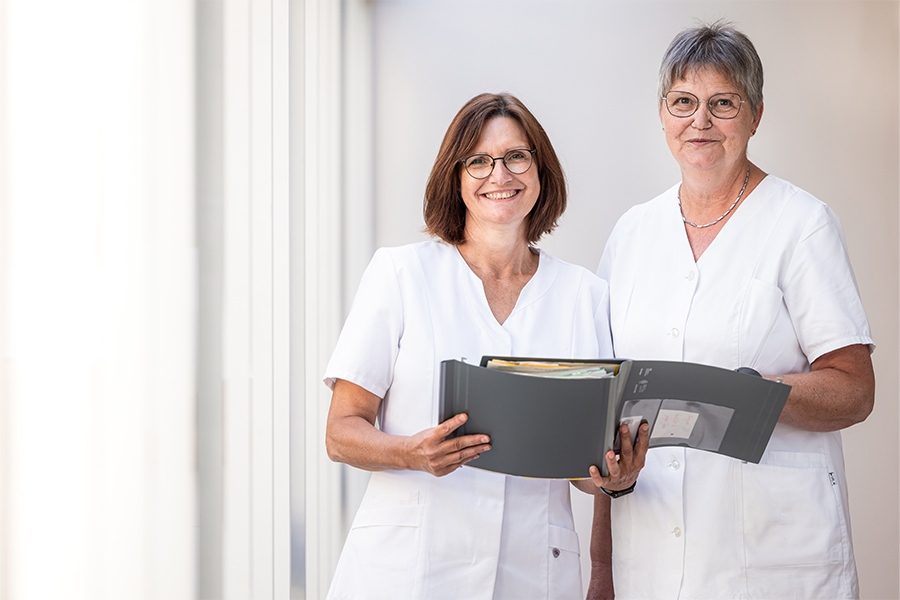 Zwei Frauen vom Klinikpersonal halten eine Patientenakte und lächeln in die Kamera.