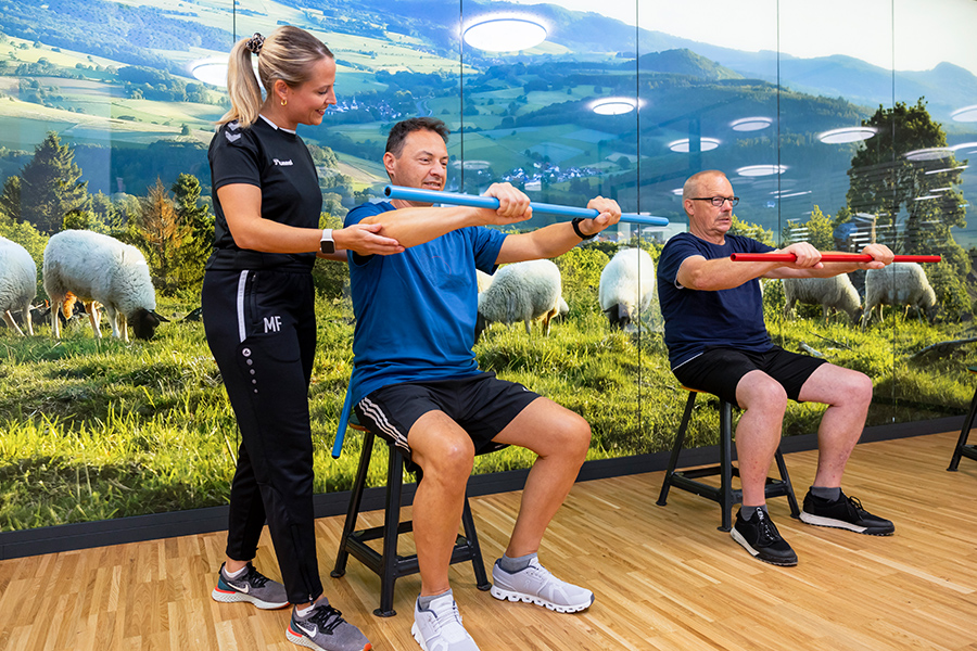 Zwei Männer in Sportbekleidung sitzen auf Hockern vor einer Fototapete mit Landschaft und Schafen und strecken ihre Hände, in denen sie einen Stab halten, nach vorne. eine Physiotherapeutin leitet sie an. Zwei Männer in Sportbekleidung sitzen auf Hockern vor einer Fototapete mit Landschaft und Schafen und strecken ihre Hände, in denen sie einen Stab halten, nach vorne. eine Physiotherapeutin leitet sie an.
