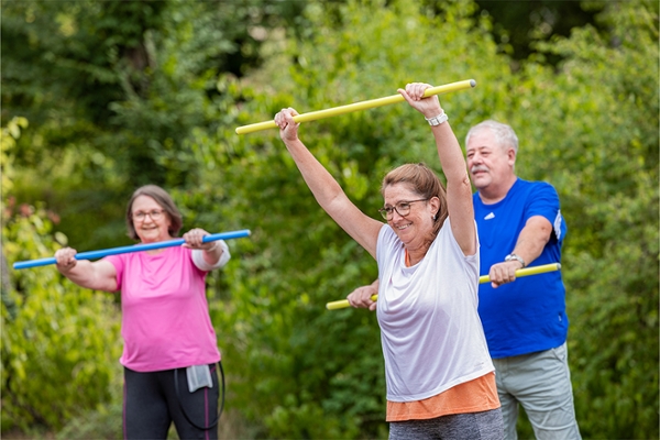 Drei Patienten machen Sportübungen mit einem Turnstab im Freien.