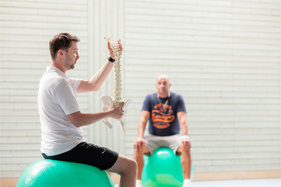 Auf Gymnastikbällen sitzend erklärt ein Therapeut seinen Patienten die Wirbelsäule.