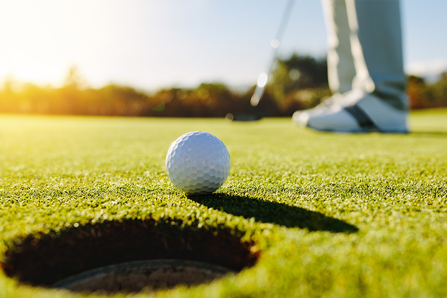 Ein Golfball liegt kurz vor dem Loch. Im Hintergrund steht eine Person mit Golfschläger.